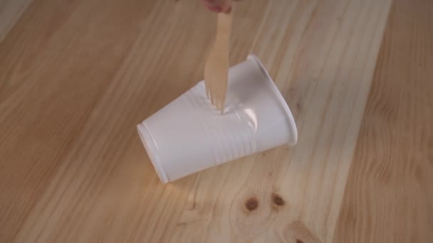 Egy környezetbarát anyagból készült villa ráncol egy fehér műanyag poharat egy fa asztalon. A biológiai ételek használata és a műanyag edények használatának elutasítása - Felvétel, videó