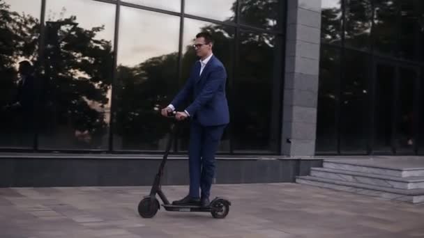Young businessman in elegant blue suit and glasses riding an electric scooter for a business meeting in the office, office buildings, business man, electric transport, ecological transport. Side view - Imágenes, Vídeo