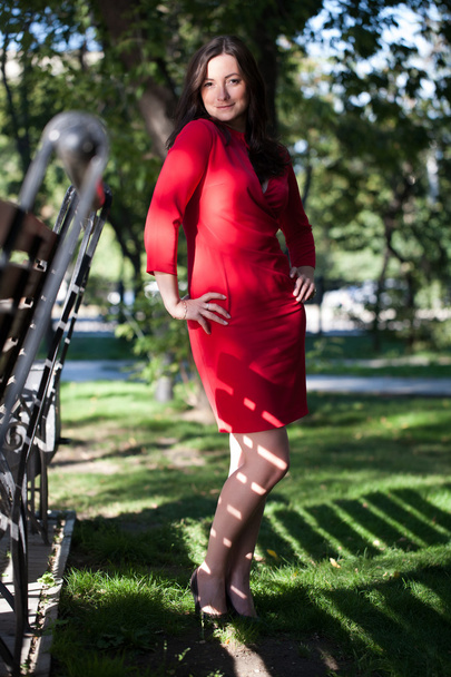 Ragazza in abito rosso è in piedi in un parco vicino alla panchina
 - Foto, immagini