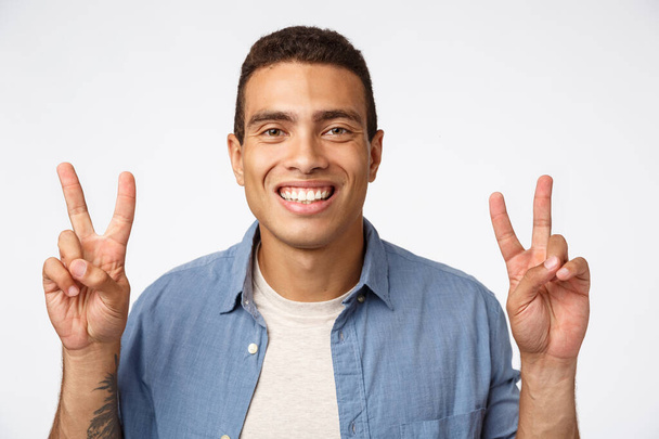 Cheerful young athletic man in casual outfit smiling happily, showing peace signs or quotation, standing carefree white background, play secret santa, ready bring joy coworkers holiday - Photo, Image