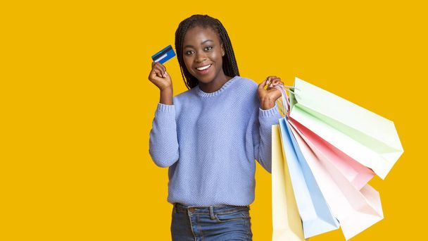 Happy african girl recommending credit card, holding shopping bags - 写真・画像
