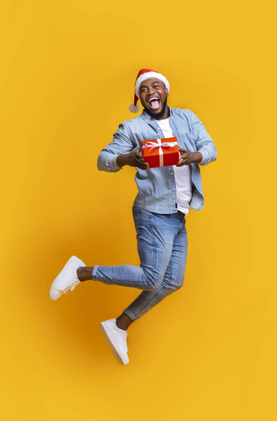 African american man in santa hat jumping with gift box - Фото, изображение