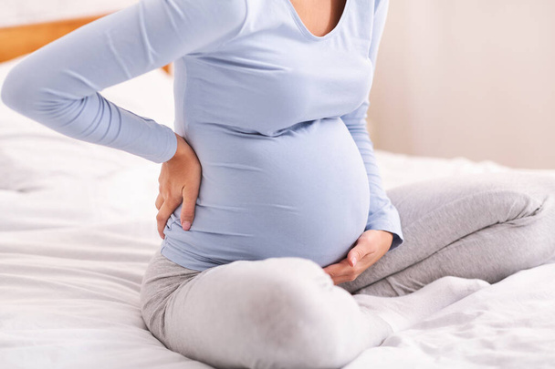 Zwangere dame met rugpijn zittend op bed in de slaapkamer, betast - Foto, afbeelding