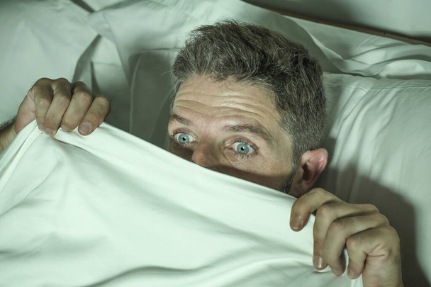  gestresste en bange man alleen in bed 's nachts wakker in angst na een nachtmerrie gevoel paranoïde houden van de deken in grappige paniek gezicht uitdrukking  - Foto, afbeelding