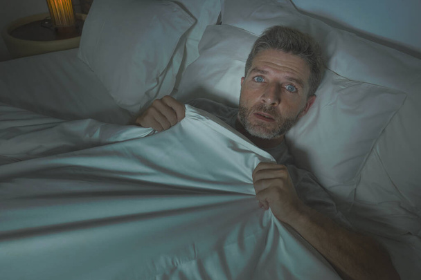  stressed and scared man alone in bed awake at night in fear after having a nightmare feeling paranoid holding the blanket in funny panic face expression  - Photo, Image