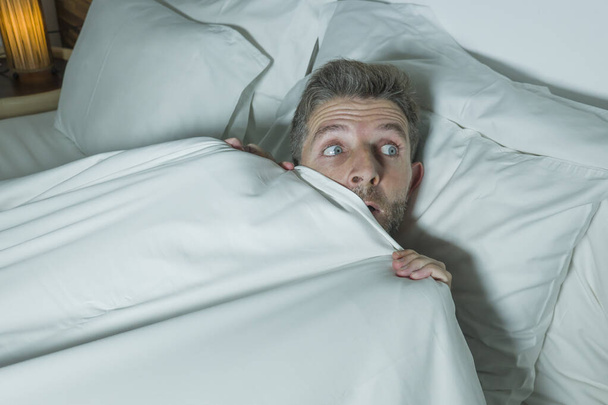  stressed and scared man alone in bed awake at night in fear after having a nightmare feeling paranoid holding the blanket in funny panic face expression  - Φωτογραφία, εικόνα