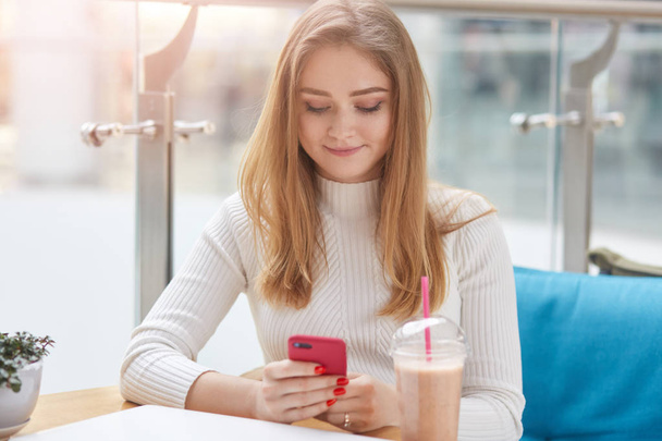 entzücktes positives Mädchen, das sein rotes Smartphone in der Hand hält, zärtlich lächelt, soziale Netzwerke nutzt, allein im Café sitzt, Coctail auf dem Tisch hat, gut gelaunt ist. Tech-Konzept. - Foto, Bild