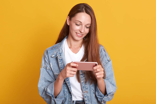 Image of tender attractive girl smiling sincerely, holding pink smartphone in her hands, looking at its screen, watching video, being fond of new technologies, wearing trendy clothes. Tech concept. - Foto, immagini