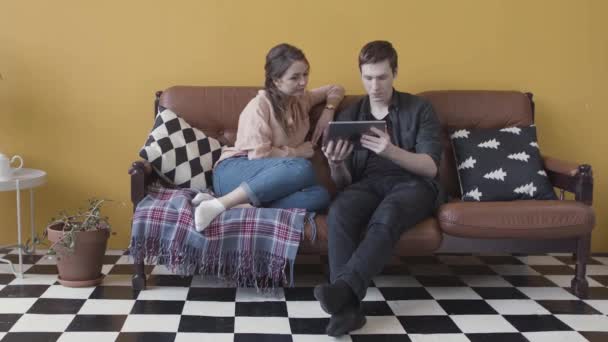 Νεαρό χαμογελαστό ζευγάρι με ψηφιακό tablet χαλαρώνοντας στον καναπέ στο σπίτι στο κομψό δωμάτιο. Στικ. Άνδρας και γυναίκα περιηγούνται μαζί στη συσκευή τους. - Πλάνα, βίντεο