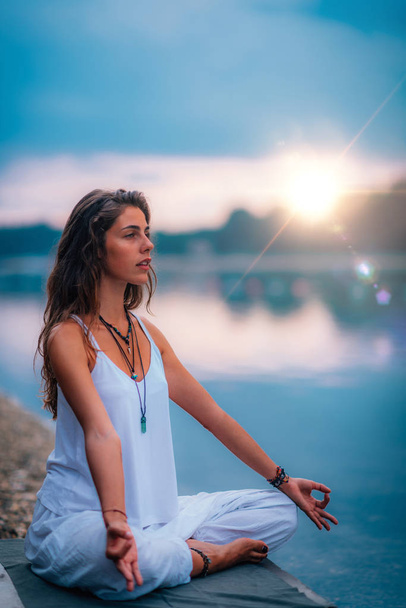 Mooie jonge vrouw die yoga oefeningen doet bij het meer. Zittend in lotuspositie - Foto, afbeelding
