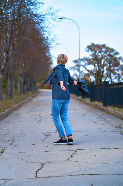 Happy blond woman walking and spinning through autumn city park, dressed in blue jeans and jacket - Photo, Image