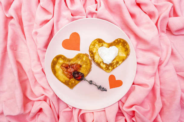 Heart shaped pancakes for romantic breakfast with strawberry jam, silver spoon and paper hearts. Shrovetide (carnival) concept. On pink cloth linen background, top view - Photo, Image