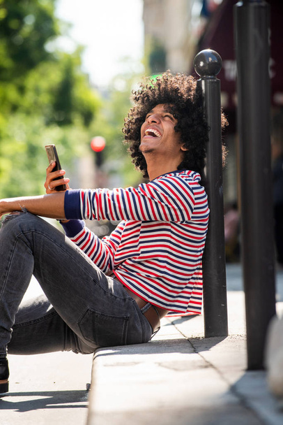 Portret szczęśliwego młodzieńca siedzącego na ulicy z telefonem komórkowym i śmiejącego się patrząc w górę - Zdjęcie, obraz
