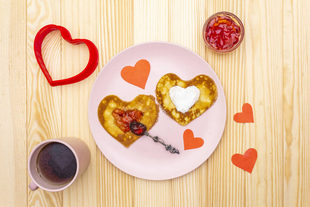 Naleśniki w kształcie serca na romantyczne śniadanie z dżemem truskawkowym i czarną herbatą. Koncepcja shrovetide (karnawał). Na drewnianym tle, widok z góry - Zdjęcie, obraz