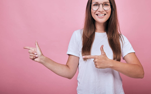 Joyful girl in T-shirt and round glasses shows forefingers to side - Foto, Imagen