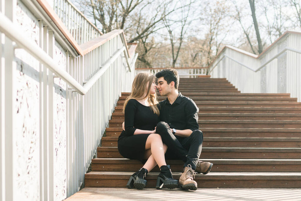 Όμορφο νεαρό πολυφυλετικό ζευγάρι, ερωτευμένο φοιτητικό ζευγάρι, κάθεται σε ξύλινη σκάλα στην πόλη. Όμορφος Τούρκος μελαχρινός αγκαλιάζει λευκή με μαύρα ρούχα. Ημερομηνία νέοι άνθρωποι σε ηλιόλουστο καιρό - Φωτογραφία, εικόνα