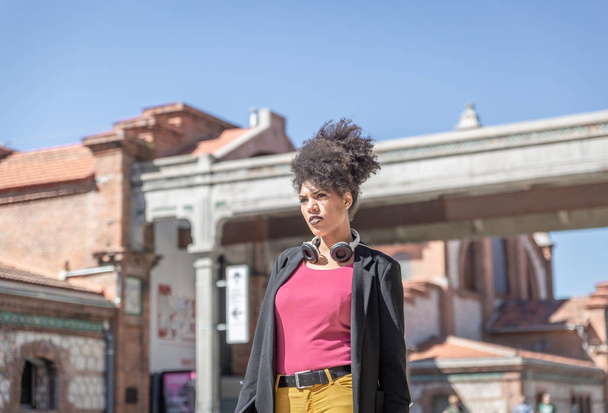 Расовая женщина с проникающим взглядом, в черной куртке и наушниках вокруг шеи, идет по городской улице в солнечный день
 - Фото, изображение