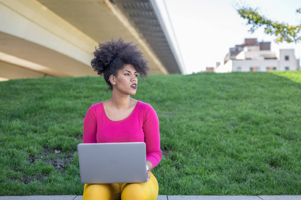 Расова дівчина з афро волоссям, дивиться на неї ліворуч задумливо, сидячи в тихому парку, що працює зі своїм ноутбуком
 - Фото, зображення