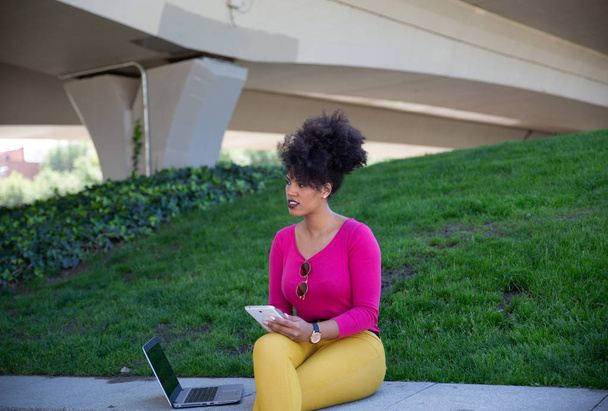attraktive brünette Unternehmerin mit Afro-Haaren, arbeitet mit Tablet und Laptop in einem entspannten Park am Stadtrand - Foto, Bild