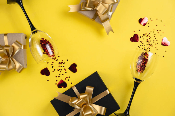 Valentine's Day background. Two clinking wine glasses, various handmade gift boxes decorated with red heart confetti on yellow background. Valentines day concept. Flat lay, top view, copy space - Fotoğraf, Görsel
