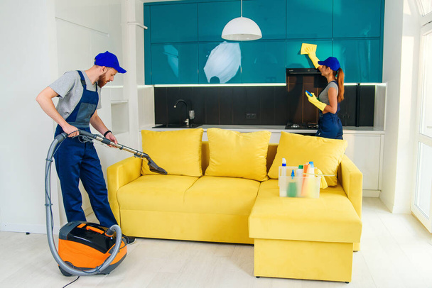 Young man with vacuum cleaner washing with vapor yellow sofa and beautiful woman wipes cuisine furniture. - Photo, Image