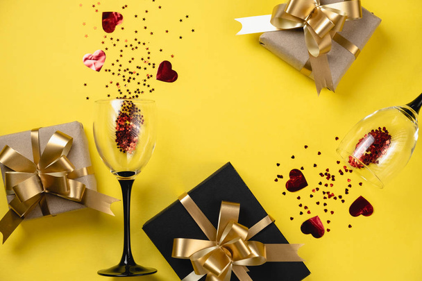 Valentine's Day background. Two clinking wine glasses, various handmade gift boxes decorated with red heart confetti on yellow background. Valentines day concept. Flat lay, top view, copy space - Φωτογραφία, εικόνα