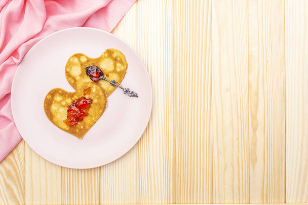 herzförmige Pfannkuchen zum romantischen Frühstück mit Erdbeermarmelade und Silberlöffel. Fastnachtskonzept. auf Holzgrund mit rosa Leinentuch, Draufsicht - Foto, Bild