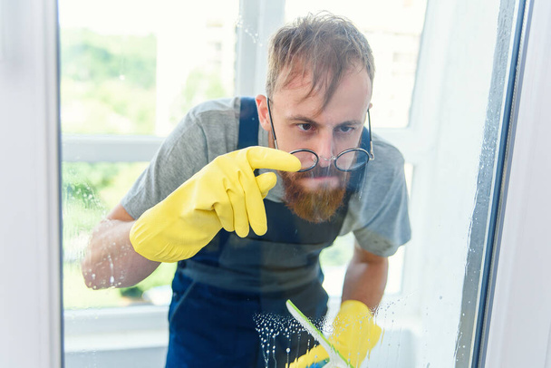 Cheerful bearded man in funny eyeglasses cleans the window with detergents and remove dirt with his finger in gloves. - Foto, Imagen