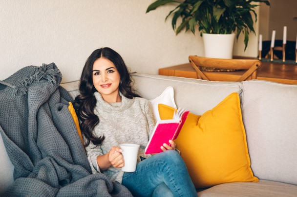Jeune belle femme reposant sur un canapé, tenant une tasse de thé ou de café, lisant un livre
 - Photo, image