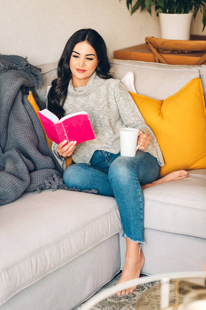 Mladá krásná žena odpočívající na pohovce, drží šálek čaje nebo kávy, čte knihu - Fotografie, Obrázek