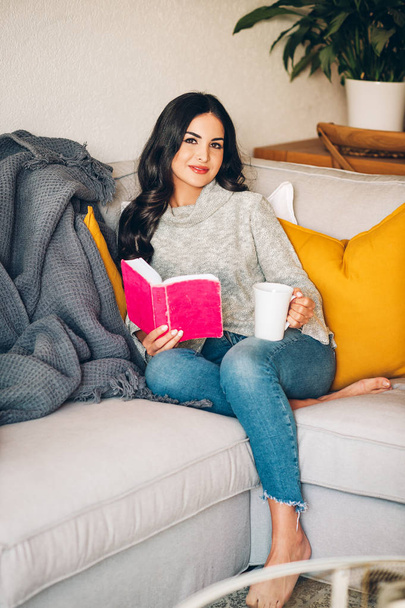 Joven mujer hermosa descansando en un sofá, sosteniendo una taza de té o café, leyendo un libro
 - Foto, imagen