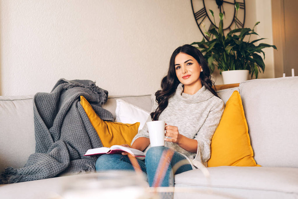 Genç, güzel bir kadın bir kanepede dinleniyor, elinde çay ya da kahve tutuyor, kitap okuyor. - Fotoğraf, Görsel