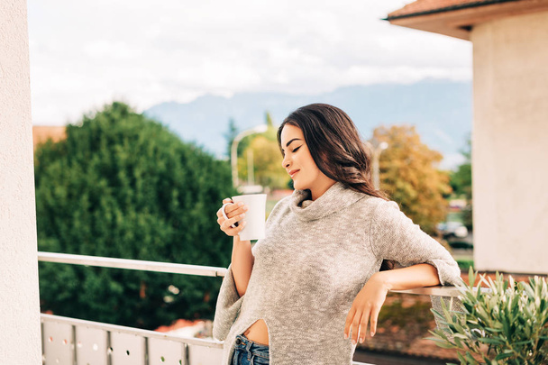 Romantic portrait of beautiful young woman wearing grey pullover, relaxing on the balcony, holding cup of tea or coffee - Foto, Imagen