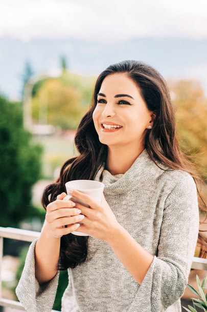 Романтичний портрет красивої молодої жінки в сірому пуловері, що розслабляється на балконі, тримає чашку чаю або кави
 - Фото, зображення