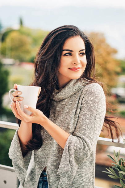 Romantisch portret van een mooie jonge vrouw met een grijze trui, ontspannen op het balkon, met een kopje thee of koffie - Foto, afbeelding