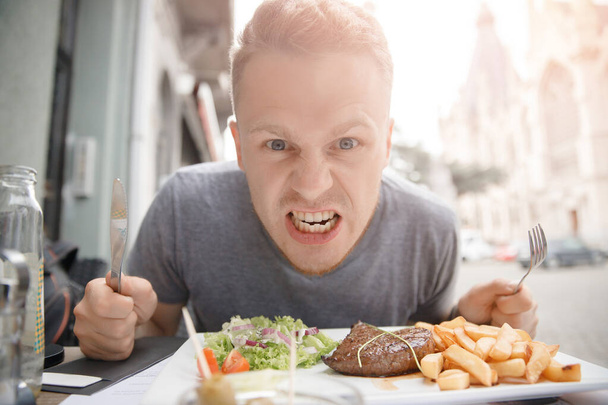 Very hungry man prepares to eat juicy steak and French fries, holds knife and fork - Photo, Image