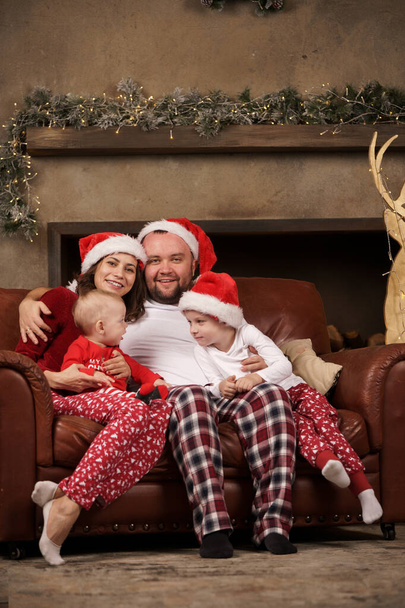 Photo of young parents in Santas caps and sons on leather sofa in studio - Photo, Image
