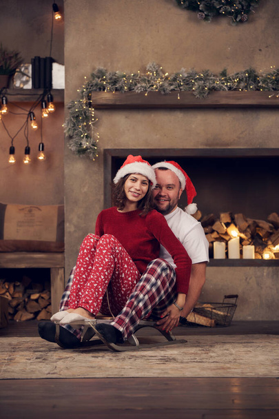 Foto de pareja feliz en gorras de Santas sentados en trineo en la habitación
 - Foto, Imagen
