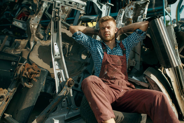 Reparador masculino relaxando no ferro-velho do carro no dia de verão. Sucata de automóveis, sucata de veículos, lixo de automóveis, transporte abandonado, danificado e esmagado
 - Foto, Imagem