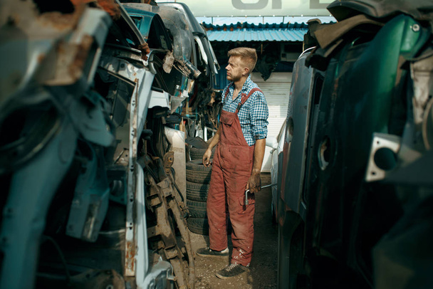 Dirty male repairman choosing spare parts on car junkyard. Auto scrap, vehicle junk, automobile garbage, abandoned, damaged and crushed transport - 写真・画像