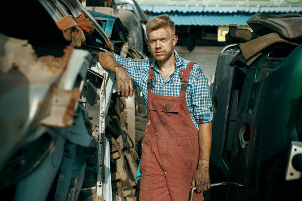 Male repairman poses on car junkyard. Auto scrap, vehicle junk, automobile garbage, abandoned, damaged and crushed transport - 写真・画像