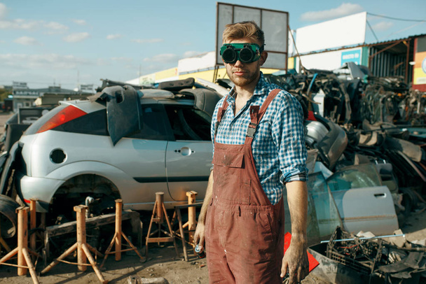 Male repairman in welding glasses on car scrapyard. Auto scrap, vehicle junk, automobile garbage. Abandoned, damaged and crushed transport, junkyard - 写真・画像