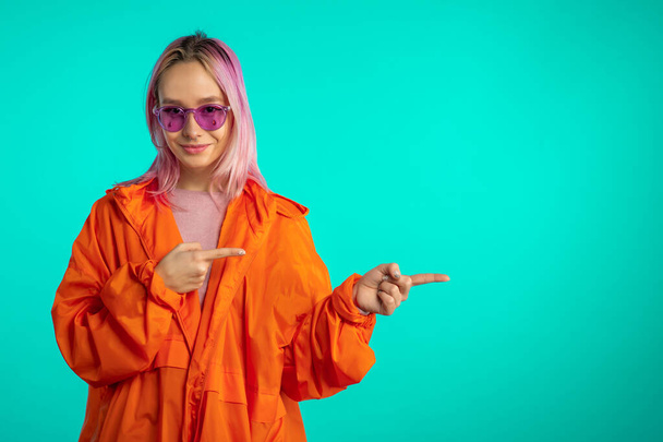  Portrait of girl with pink hair, she indicates with arms on her left with copy space. Happy smiling woman presenting and showing something isolated on blue background. - Valokuva, kuva