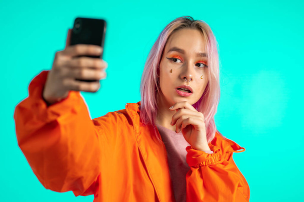 Girl with pink hair and extraordinary appearance make selfie on blue background. Using modern technology - smartphone, social networks - Фото, зображення