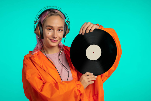 Pretty girl with pink hair enjoys listening to music. Hipster woman with extraordinary appearance in orange raincoat holding vinyl record.on blue background. - Photo, image