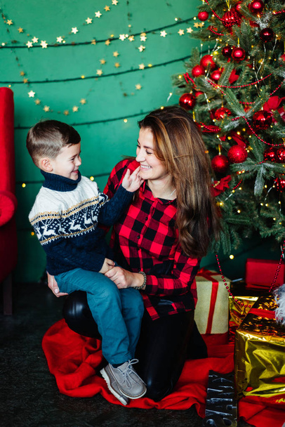 Mama z synem w swetrze świątecznym na drutach raduje się z prezentów, siedząc przy drzewie noworocznym. Wesołych Świąt z prezentami. Zimowy nastrój - Zdjęcie, obraz