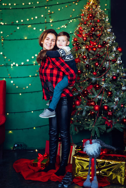Мама с сыном в вязаном рождественском свитере радуется подаркам, сидя рядом с новогодней елкой. Счастливого Рождества с подарками. Зимнее настроение
 - Фото, изображение