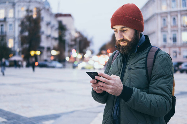 Молодой бородатый мужчина смотрит на экран мобильного телефона во время прогулки по улице
 - Фото, изображение
