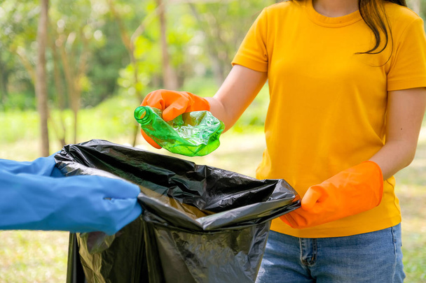 Hands of yellow t-shirt woman collect the green bottle as garbage to black bag with someone help to hold it. - 写真・画像