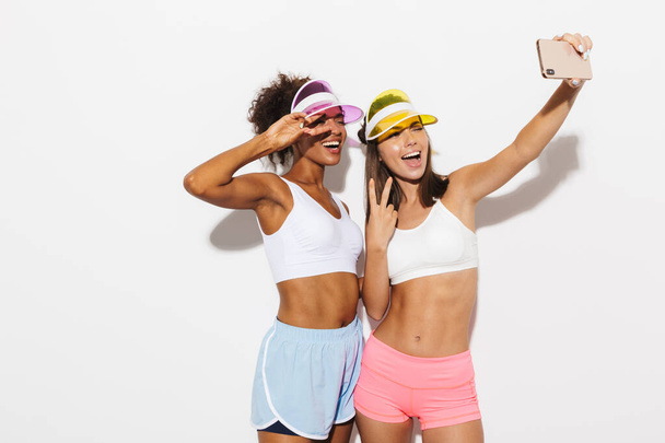 Portrait of two cheerful multinational women wearing visor hats taking selfie photo on cellphone and gesturing peace sign - Fotó, kép